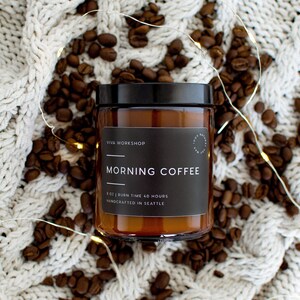 May include: A brown glass candle jar with a black label that reads "Viva Workshop" and "Morning Coffee". The label also states "8 oz | Burn Time 40 Hours | Handcrafted in Seattle". The candle is surrounded by coffee beans and white string lights.