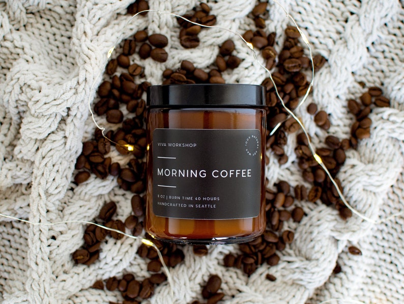 May include: A brown glass candle jar with a black label that reads "Viva Workshop" and "Morning Coffee". The label also states "8 oz | Burn Time 40 Hours | Handcrafted in Seattle". The candle is surrounded by coffee beans and white string lights.