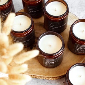 Puede incluir: Una colecci&oacute;n de velas en frasco de vidrio &aacute;mbar con cera blanca y etiquetas negras con el texto Viva Workshop, Cranberry Prosecco, Pumpkin Spice, Fallen Leaves, Morning Coffee, Leather & Smoke y Oakmoss & Amber sobre un disco de madera.
