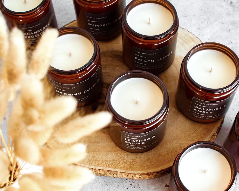 Puede incluir: Una colecci&oacute;n de velas en frasco de vidrio &aacute;mbar con cera blanca y etiquetas negras con el texto Viva Workshop, Cranberry Prosecco, Pumpkin Spice, Fallen Leaves, Morning Coffee, Leather & Smoke y Oakmoss & Amber sobre un disco de madera.