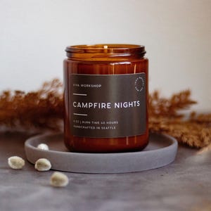 May include: A brown glass candle with a black label that reads "Viva Workshop Campfire Nights 8 oz | Burn Time 40 Hours Handcrafted in Seattle". The candle is sitting on a gray coaster.