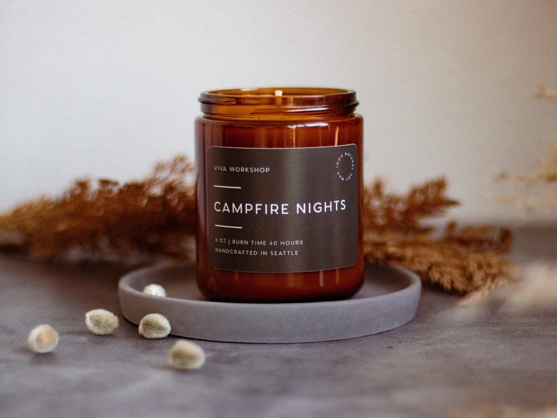 May include: A brown glass candle with a black label that reads "Viva Workshop Campfire Nights 8 oz | Burn Time 40 Hours Handcrafted in Seattle". The candle is sitting on a gray coaster.