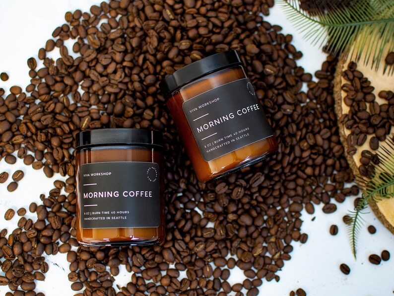May include: Two amber glass candle jars with black lids and labels. The labels read "Viva Workshop" and "Morning Coffee". The candles are surrounded by coffee beans.