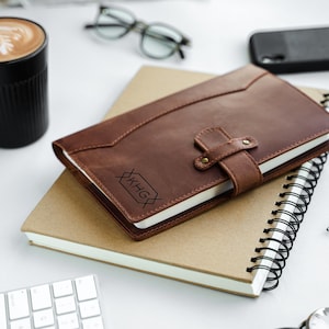 Peut inclure: Un journal en cuir marron avec une fermeture &agrave; lani&egrave;re et un monogramme en creux repose sur un cahier &agrave; spirales. Une tasse noire avec un latte, des lunettes et un t&eacute;l&eacute;phone sont &eacute;galement sur la surface blanche.