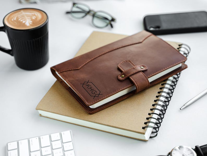 Peut inclure: Un journal en cuir marron avec une fermeture &agrave; lani&egrave;re et un monogramme en creux repose sur un cahier &agrave; spirales. Une tasse noire avec un latte, des lunettes et un t&eacute;l&eacute;phone sont &eacute;galement sur la surface blanche.