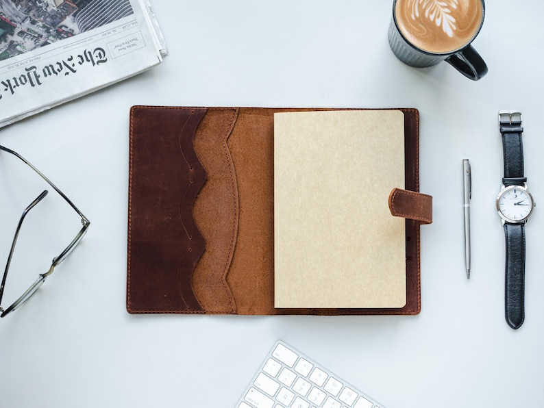 Peut inclure: Un journal en cuir marron ouvert avec une page vierge, un stylo et une montre-bracelet avec un bracelet noir. Une tasse de caf&eacute; et un journal sont &eacute;galement visibles, sugg&eacute;rant un espace de travail ou un bureau.