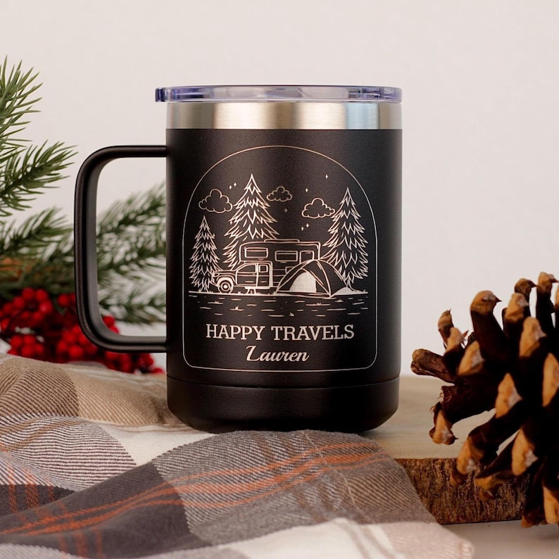 Puede incluir: Taza de viaje negra con borde y asa plateados. La taza presenta un gr&aacute;fico blanco de una caravana, una tienda de campa&ntilde;a y &aacute;rboles bajo las palabras "Happy Travels" y un nombre. La taza tiene una tapa de pl&aacute;stico transparente.