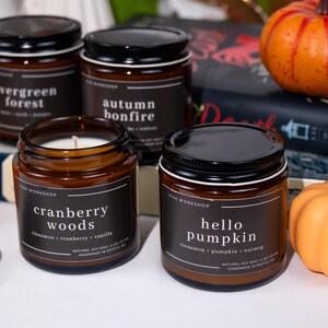 May include: Four amber glass candle jars with black lids and white labels. The candles are arranged on a white surface with two decorative pumpkins. The labels read "evergreen forest", "autumn bonfire", "cranberry woods", and "hello pumpkin".