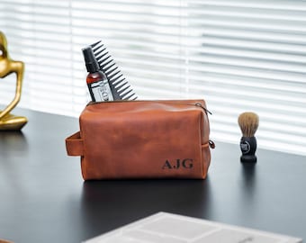 May include: A brown leather toiletry bag with a zipper closure and a handle. The bag is monogrammed with the initials "AJG". The bag is filled with a comb and a bottle of beard oil.