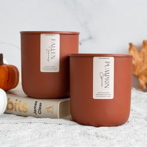 Puede incluir: Dos velas de cera de soja marrones con etiquetas blancas. La vela m&aacute;s grande est&aacute; etiquetada como "Fallen Leaves" y la vela m&aacute;s peque&ntilde;a est&aacute; etiquetada como "Pumpkin Spice". Las velas est&aacute;n sobre un libro y una superficie blanca y peluda con calabazas blancas y naranjas.