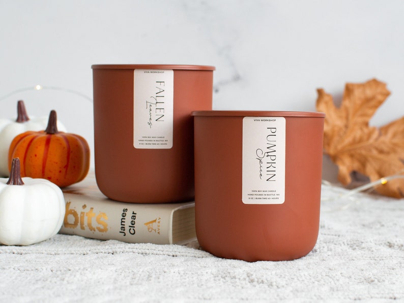 Puede incluir: Dos velas de cera de soja marrones con etiquetas blancas. La vela m&aacute;s grande est&aacute; etiquetada como "Fallen Leaves" y la vela m&aacute;s peque&ntilde;a est&aacute; etiquetada como "Pumpkin Spice". Las velas est&aacute;n sobre un libro y una superficie blanca y peluda con calabazas blancas y naranjas.