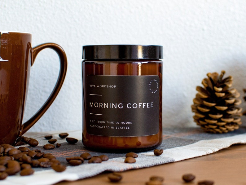 May include: A brown glass candle jar with a black label that reads "Viva Workshop Morning Coffee 8 oz | Burn Time 40 Hours Handcrafted in Seattle". The candle is sitting on a white and brown plaid fabric with coffee beans scattered around it.