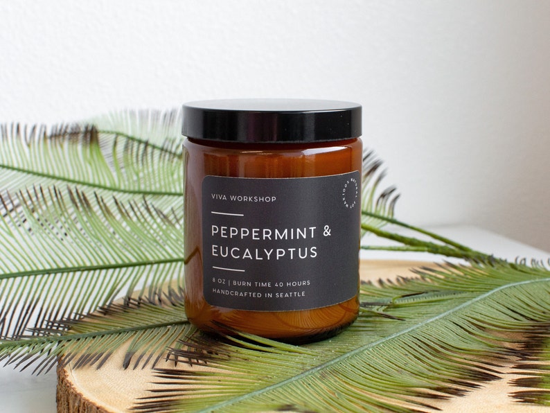 Puede incluir: Un tarro de vidrio marr&oacute;n con una vela, una tapa negra y una etiqueta. La etiqueta dice "Viva Workshop", "Peppermint & Eucalyptus", "8 oz | Burn Time 40 Hours", y "Handcrafted in Seattle".