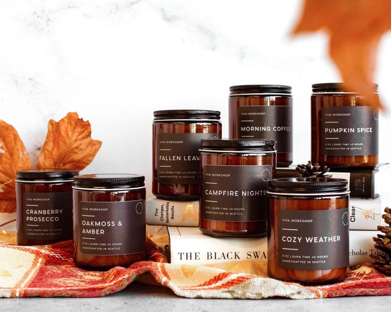 Puede incluir: Una colecci&oacute;n de seis velas en frasco de vidrio &aacute;mbar con tapas y etiquetas negras. Las etiquetas presentan el texto Viva Workshop y nombres de aromas como Cranberry Prosecco, Oakmoss & Amber, Fallen Leaves, Morning Coffee, Campfire Nights, Pumpkin Spice y Cozy Weather.