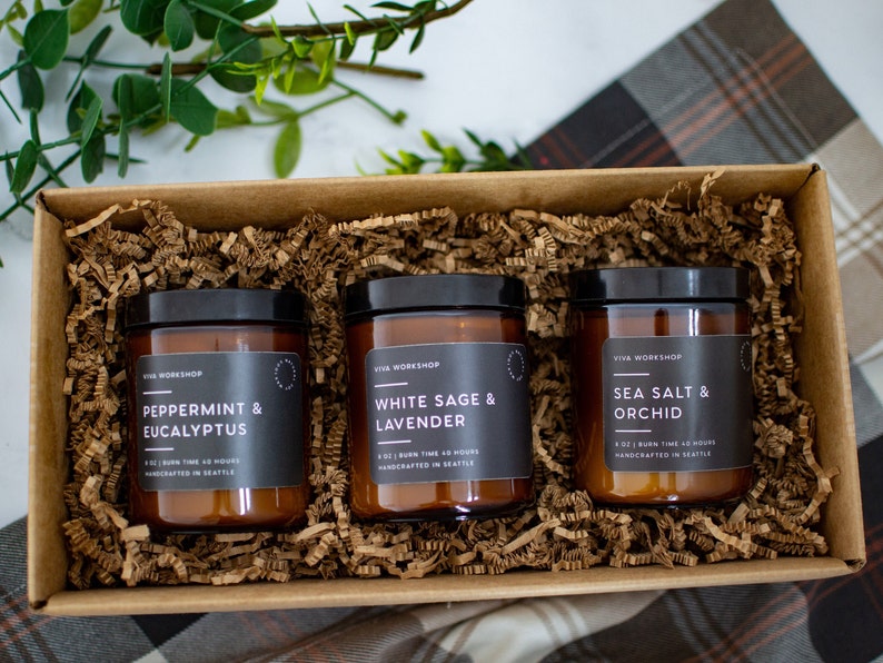 Puede incluir: Una caja de regalo llena de virutas de papel marr&oacute;n que contiene tres tarros de velas de vidrio &aacute;mbar con etiquetas negras. Las etiquetas dicen "Peppermint & Eucalyptus", "White Sage & Lavender" y "Sea Salt & Orchid". Cada vela tiene un tiempo de combusti&oacute;n de 40 horas y est&aacute; hecha a mano en Seattle.