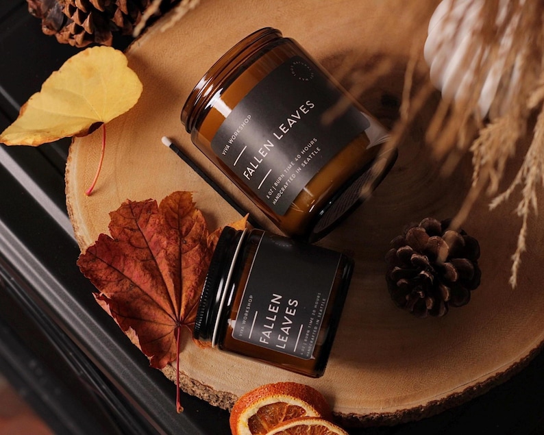 Puede incluir: Dos velas en frasco de vidrio &aacute;mbar con etiquetas negras que dicen FALLEN LEAVES, 8 OZ | BURN TIME 40 HOURS, HANDCRAFTED IN SEATTLE. Las velas est&aacute;n rodeadas de hojas de oto&ntilde;o secas, una pi&ntilde;a de pino y rodajas de naranja secas.