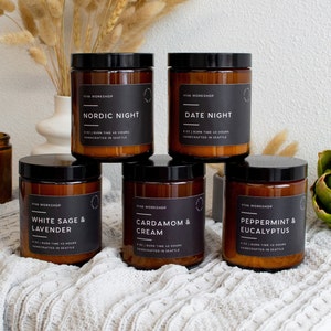 May include: Six amber glass candle jars with black lids and white labels. The candles are arranged in two rows of three. The labels have the names of the scents: Nordic Night, Date Night, White Sage & Lavender, Cardamom & Cream, Peppermint & Eucalyptus. The candles are handcrafted in Seattle.
