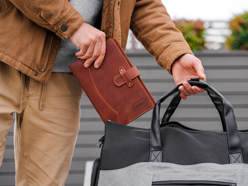 May include: A brown leather portfolio with a buckle closure and the initials "KHG" embossed on the front. The portfolio is held next to a black and gray duffel bag. The person is wearing a brown jacket and khaki pants.