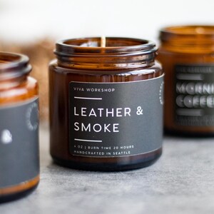 Puede incluir: Tres velas en frascos de vidrio &aacute;mbar con etiquetas negras. La etiqueta de la vela central dice VIVA WORKSHOP LEATHER & SMOKE 6 OZ | BURN TIME 20 HOURS HANDCRAFTED IN SEATTLE. Las velas presentan una mecha visible y un dise&ntilde;o cil&iacute;ndrico.