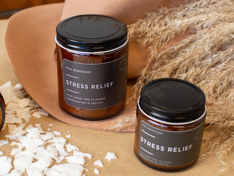 May include: Two amber glass candle jars with black lids and labels that read "Viva Workshop" and "Stress Relief". The candles are 8 oz and have a burn time of 40 hours. They are handcrafted in Seattle.