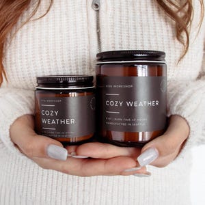 Puede incluir: Dos velas en frascos de vidrio &aacute;mbar con tapas negras de rosca y etiquetas negras. Las etiquetas muestran el texto VIVA WORKSHOP y COZY WEATHER. El frasco peque&ntilde;o indica 4 oz y 20 horas de combusti&oacute;n, el grande indica 8 oz y 40 horas.
