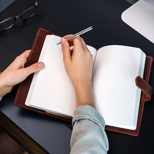 Peut inclure: Un journal en cuir marron avec des pages lign&eacute;es est ouvert, tenu par une personne. Un stylo argent&eacute; avec une pointe bleue est pos&eacute; sur le papier, pr&ecirc;t &agrave; &eacute;crire. Le journal a une fermeture &agrave; lani&egrave;re en cuir. Lunettes et ordinateur en arri&egrave;re-plan.
