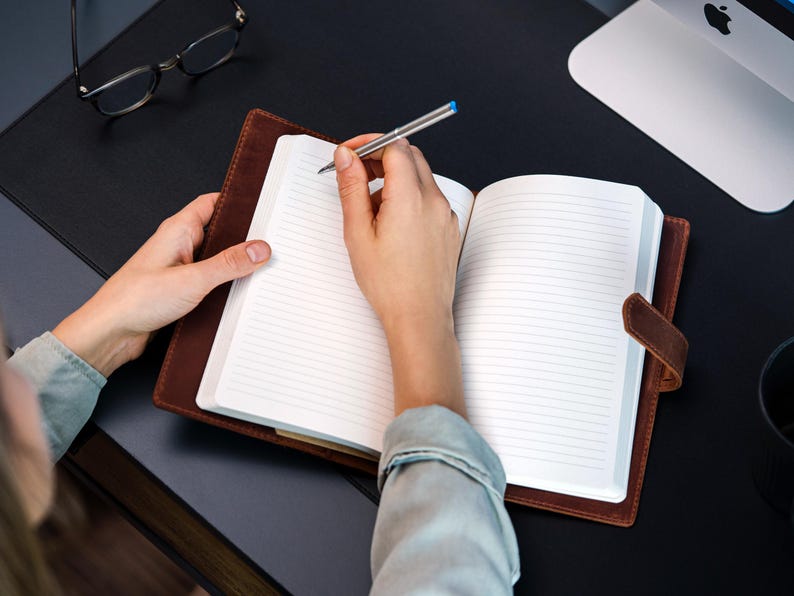 Peut inclure: Un journal en cuir marron avec des pages lign&eacute;es est ouvert, tenu par une personne. Un stylo argent&eacute; avec une pointe bleue est pos&eacute; sur le papier, pr&ecirc;t &agrave; &eacute;crire. Le journal a une fermeture &agrave; lani&egrave;re en cuir. Lunettes et ordinateur en arri&egrave;re-plan.