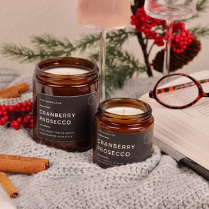 Puede incluir: Dos velas en frasco de vidrio &aacute;mbar con etiquetas negras que dicen Cranberry Prosecco, 8 oz, tiempo de combusti&oacute;n 40 horas, hechas a mano en Seattle. Las velas est&aacute;n sobre una tela de punto gris junto a ramas de canela y un libro.