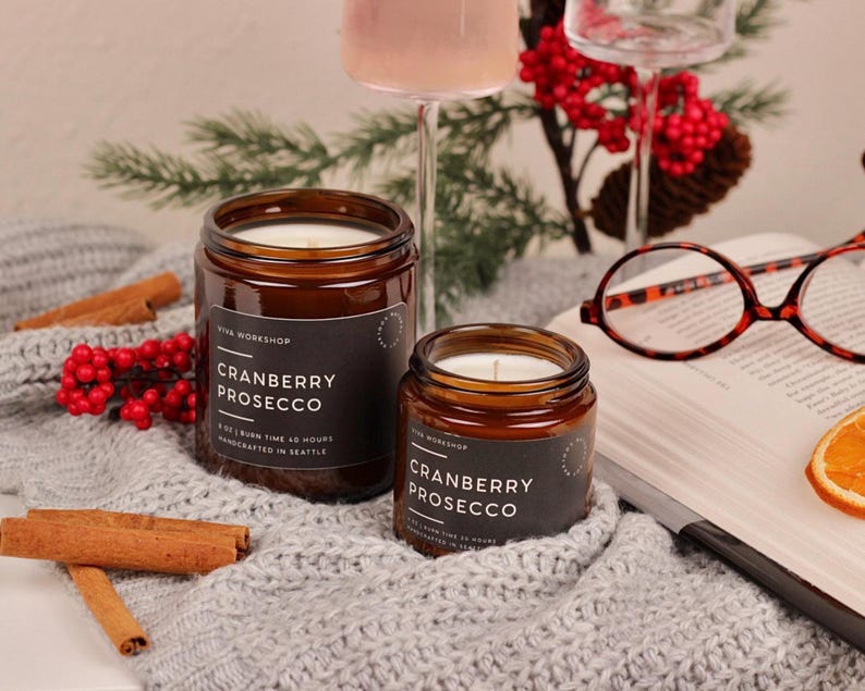 Puede incluir: Dos velas en frasco de vidrio &aacute;mbar con etiquetas negras que dicen Cranberry Prosecco, 8 oz, tiempo de combusti&oacute;n 40 horas, hechas a mano en Seattle. Las velas est&aacute;n sobre una tela de punto gris junto a ramas de canela y un libro.