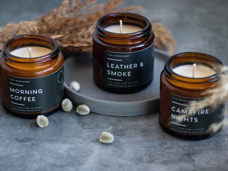 May include: Three amber glass candle jars with black labels. The candles are labeled "Morning Coffee", "Leather & Smoke", and "Campfire Nights".