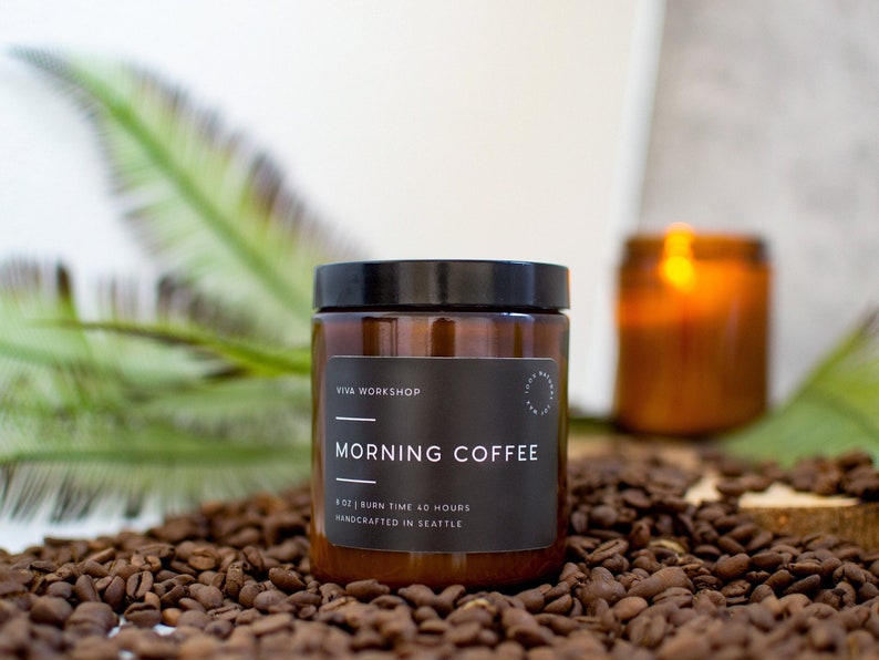 May include: A brown glass candle jar with a black label that reads "Viva Workshop Morning Coffee 4 oz | Burn Time 40 Hours Handcrafted in Seattle". The candle is sitting on a bed of coffee beans.