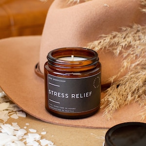 May include: A brown glass candle jar with a black label that reads "STRESS RELIEF" and "Viva Workshop". The candle is lit and the jar is sitting on a brown surface.
