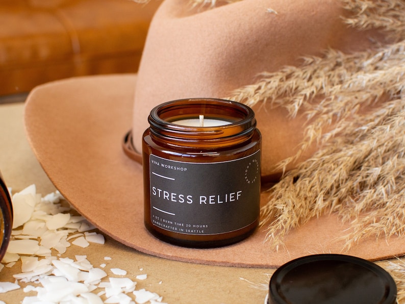 May include: A brown glass candle jar with a black label that reads "STRESS RELIEF" and "Viva Workshop". The candle is lit and the jar is sitting on a brown surface.