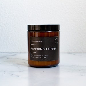 May include: A brown glass candle jar with a black lid and a white label that reads "Viva Workshop" and "Morning Coffee". The label also states "8 oz | Burn Time 40 Hours" and "Handcrafted in Seattle".