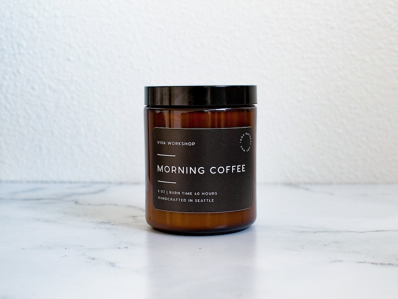 May include: A brown glass candle jar with a black lid and a white label that reads "Viva Workshop" and "Morning Coffee". The label also states "8 oz | Burn Time 40 Hours" and "Handcrafted in Seattle".