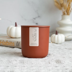 Puede incluir: Una vela marr&oacute;n con una etiqueta blanca que dice "Pumpkin Spice" en blanco, en fuente sans-serif. La vela est&aacute; sobre una superficie blanca y peluda.