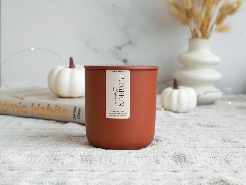 Puede incluir: Una vela marr&oacute;n con una etiqueta blanca que dice "Pumpkin Spice" en blanco, en fuente sans-serif. La vela est&aacute; sobre una superficie blanca y peluda.