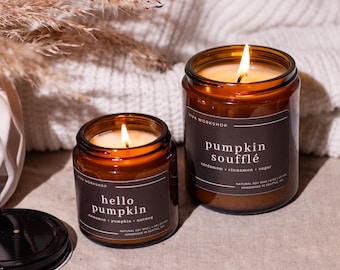 May include: Two lit soy candles in amber glass jars. One reads "hello pumpkin" and the other "pumpkin soufflé." The labels list ingredients. The candles are on a neutral surface with a white knitted blanket in the background.