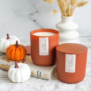 Puede incluir: Dos velas de color terracota con etiquetas que dicen "Pumpkin Spice" y "Fallen Leaves". Las velas est&aacute;n sobre un libro con tres peque&ntilde;as calabazas blancas y naranjas.