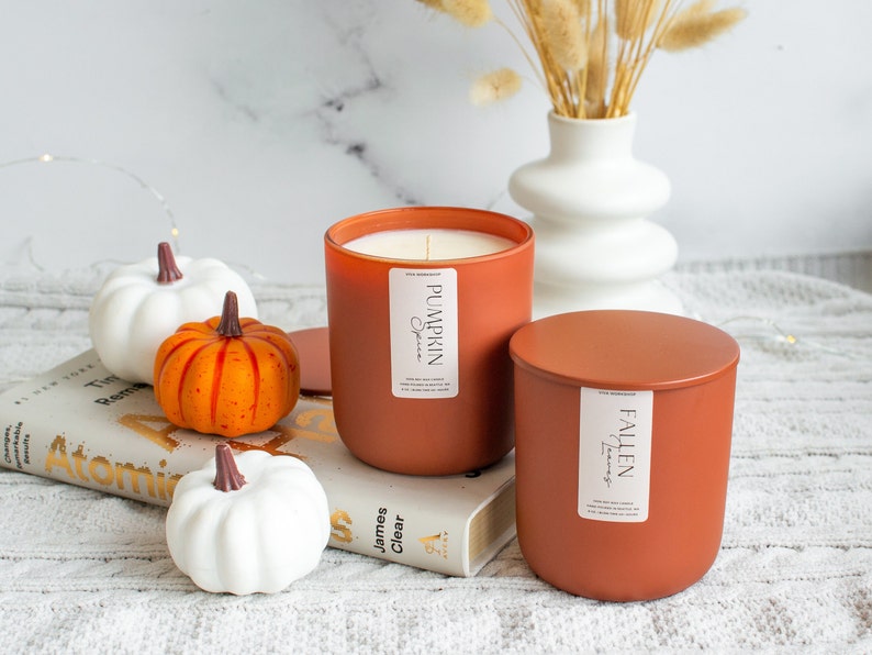 Puede incluir: Dos velas de color terracota con etiquetas que dicen "Pumpkin Spice" y "Fallen Leaves". Las velas est&aacute;n sobre un libro con tres peque&ntilde;as calabazas blancas y naranjas.