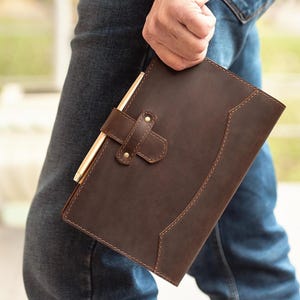 Peut inclure: Un journal en cuir marron avec un stylo fix&eacute; par une lani&egrave;re en cuir. Le journal a une bordure cousue et une fermeture &agrave; boucle. Le stylo est de couleur dor&eacute;e. Le journal est tenu par une personne portant un jean bleu.