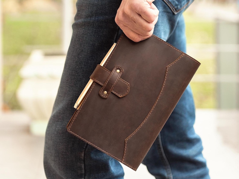 Peut inclure: Un journal en cuir marron avec un stylo fix&eacute; par une lani&egrave;re en cuir. Le journal a une bordure cousue et une fermeture &agrave; boucle. Le stylo est de couleur dor&eacute;e. Le journal est tenu par une personne portant un jean bleu.