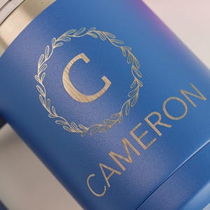 Puede incluir: Un vaso azul con el nombre "CAMERON" y la letra "C" dentro de una corona de laurel dorada. El vaso tiene un borde plateado met&aacute;lico y un acabado mate.