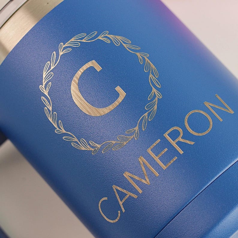 Puede incluir: Un vaso azul con el nombre "CAMERON" y la letra "C" dentro de una corona de laurel dorada. El vaso tiene un borde plateado met&aacute;lico y un acabado mate.
