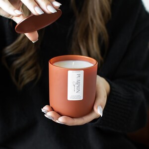 Puede incluir: Un tarro de vela de color terracota con una etiqueta blanca que dice "Pumpkin Spice". La vela est&aacute; encendida y la tapa est&aacute; siendo retirada por una mano.
