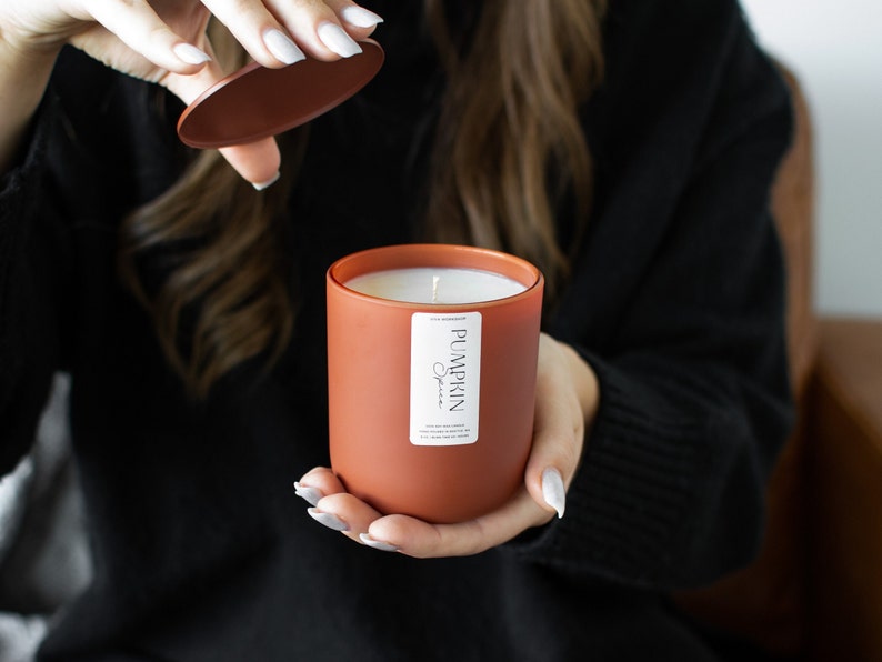 Puede incluir: Un tarro de vela de color terracota con una etiqueta blanca que dice "Pumpkin Spice". La vela est&aacute; encendida y la tapa est&aacute; siendo retirada por una mano.