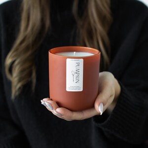 Puede incluir: Un tarro de vela de color terracota con una etiqueta blanca que dice "Pumpkin Spice". La vela est&aacute; encendida y la cera se est&aacute; derritiendo.