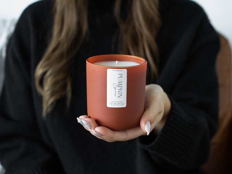 Puede incluir: Un tarro de vela de color terracota con una etiqueta blanca que dice "Pumpkin Spice". La vela est&aacute; encendida y la cera se est&aacute; derritiendo.