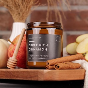 Puede incluir: Una vela en frasco de vidrio &aacute;mbar de 8 oz con una etiqueta negra que dice Apple Pie & Cinnamon, Viva Workshop y Handcrafted in Seattle. La vela est&aacute; rodeada por una manzana roja, un cuchar&oacute;n de miel, ramas de canela y una calabaza blanca.