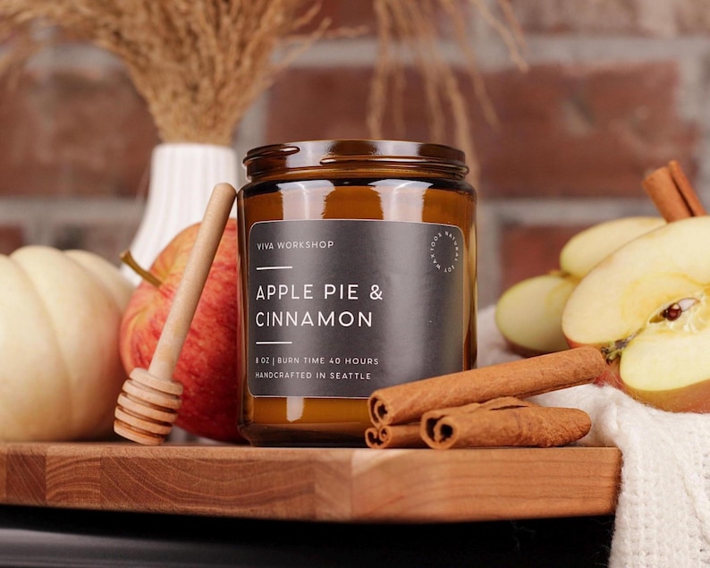 Puede incluir: Una vela en frasco de vidrio &aacute;mbar de 8 oz con una etiqueta negra que dice Apple Pie & Cinnamon, Viva Workshop y Handcrafted in Seattle. La vela est&aacute; rodeada por una manzana roja, un cuchar&oacute;n de miel, ramas de canela y una calabaza blanca.